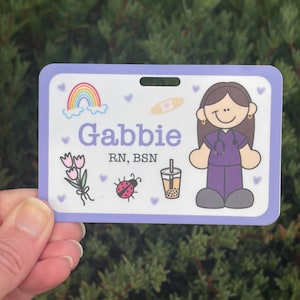 以下が含まれることがあります： 紫色の縁取りが付いた、パーソナライズされたネームバッジ。「Gabbie」という名前が紫で表示され、「RN、BSN」というタイトルも表示されます。虹、花、てんとう虫、絆創膏、バブルティーの漫画イラストが含まれています。