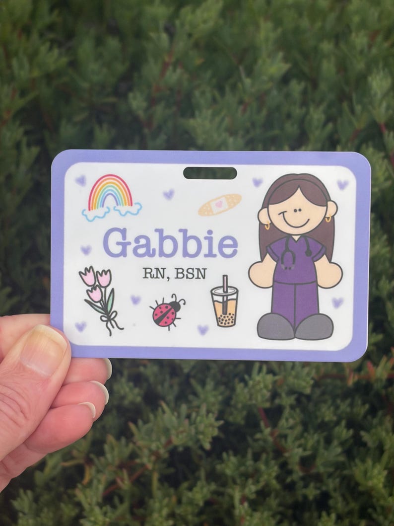 以下が含まれることがあります： 紫色の縁取りが付いた、パーソナライズされたネームバッジ。「Gabbie」という名前が紫で表示され、「RN、BSN」というタイトルも表示されます。虹、花、てんとう虫、絆創膏、バブルティーの漫画イラストが含まれています。