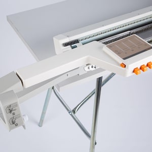 Può includere: Macchina per maglieria bianca con pannello di controllo e pulsanti arancioni. La macchina è montata su un tavolo bianco sostenuto da un supporto pieghevole argentato. La macchina ha una serie di aghi e un braccio a leva.