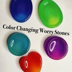 May include: Five smooth, oval and heart-shaped worry stones in blue, green, teal, purple, and red-to-orange gradient colors. The text Color Changing Worry Stones and By Mended Mind Forge is printed on the image in a black serif font.
