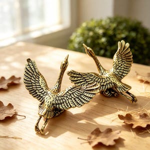 Könnte beinhalten: Zwei messingfarbene Kranichfiguren aus Metall, die im Flug mit weit ausgebreiteten Flügeln dargestellt sind. Jeder Vogel weist detaillierte, geätzte Federmuster, lange Hälse und ausgestreckte Beine auf, was eine dynamische Pose einfängt.