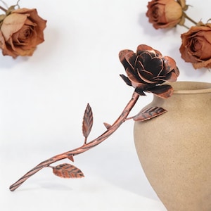 May include: A metallic rose sculpture with a dark copper finish, featuring layered petals, a textured stem with small thorns, and three distinct leaves. The sculpture is positioned at an angle, resting against the rim of a tan ceramic vase.
