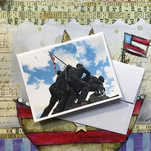 May include: A greeting card featuring a photograph of the Iwo Jima Memorial, a statue of five Marines raising the American flag on Mount Suribachi during World War II. The card has a blue sky with white clouds in the background.