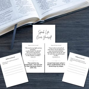 May include: An open book with a black cover and a bookmark, with several white cards featuring inspirational quotes and prompts. The cards have black text on a white background. The words "Speak Life Over Yourself" are at the top of the cards.