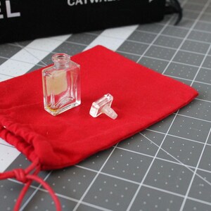 May include: A small, clear glass perfume bottle with a rectangular shape and a matching clear glass stopper, featuring a light-colored rectangular label on the front, resting on a bright red fabric drawstring pouch with matching red cords.