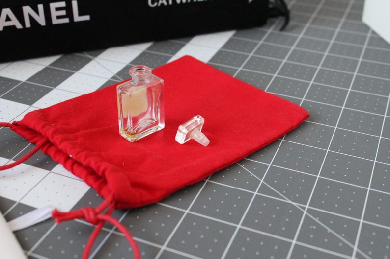 May include: A small, clear glass perfume bottle with a rectangular shape and a matching clear glass stopper, featuring a light-colored rectangular label on the front, resting on a bright red fabric drawstring pouch with matching red cords.
