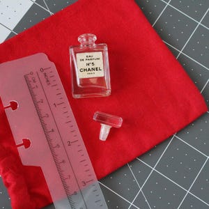 May include: A clear glass Chanel No. 5 Eau de Parfum miniature bottle with a separate glass stopper, positioned next to a translucent plastic ruler showing measurements in inches and centimeters, all resting on a red fabric pouch.
