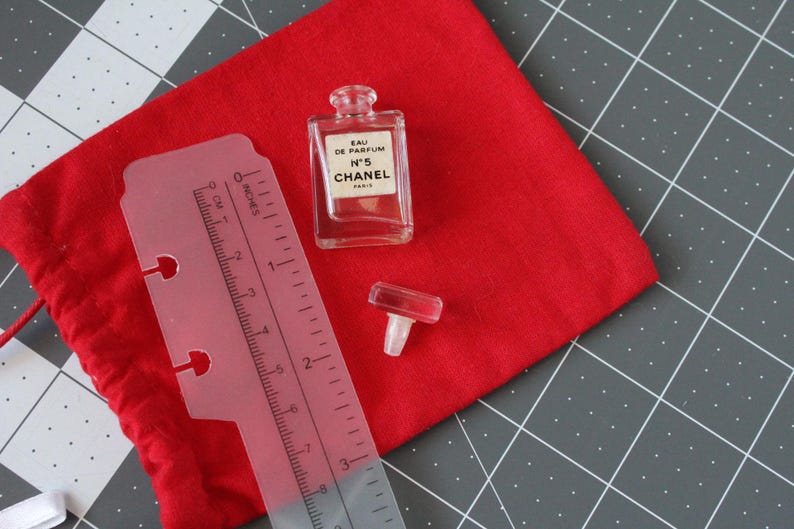 May include: A clear glass Chanel No. 5 Eau de Parfum miniature bottle with a separate glass stopper, positioned next to a translucent plastic ruler showing measurements in inches and centimeters, all resting on a red fabric pouch.