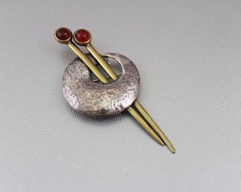 May include: A silver and brass brooch with two red stones. The brooch is shaped like a crescent moon with a hammered texture. The stones are set in the ends of two brass rods that cross each other.