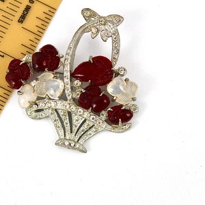 Può includere: Un broche color argento con un design a forma di cestino. Il cestino è pieno di pietre rosse e bianche. Una farfalla è in cima al cestino.