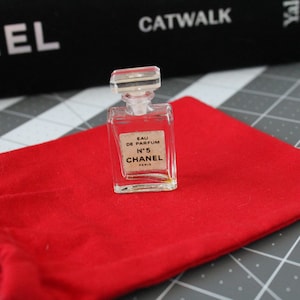 May include: A miniature clear glass perfume bottle with a rectangular stopper. The label reads EAU DE PARFUM N&deg;5 CHANEL PARIS. The bottle is positioned on a bright red fabric drawstring pouch. The glass is transparent and the liquid inside is pale.
