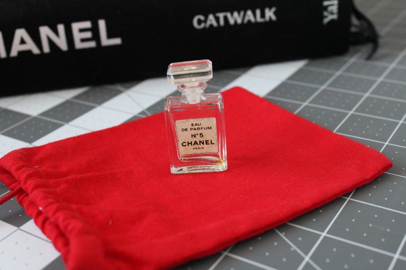 May include: A miniature clear glass perfume bottle with a rectangular stopper. The label reads EAU DE PARFUM N&deg;5 CHANEL PARIS. The bottle is positioned on a bright red fabric drawstring pouch. The glass is transparent and the liquid inside is pale.