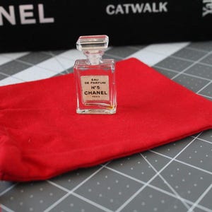 May include: A miniature clear glass perfume bottle with a rectangular stopper and a white label reading EAU DE PARFUM N&deg;5 CHANEL PARIS, resting on a bright red fabric drawstring pouch with matching red cords and a rectangular base.