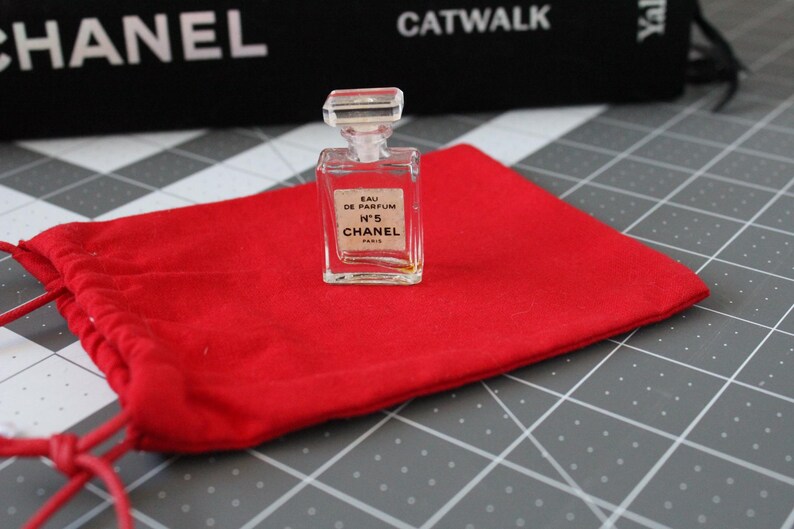 May include: A miniature clear glass perfume bottle with a rectangular stopper and a white label reading EAU DE PARFUM N&deg;5 CHANEL PARIS, resting on a bright red fabric drawstring pouch with matching red cords and a rectangular base.