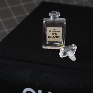 May include: A small, clear glass perfume bottle with a rectangular shape and a matching clear stopper. The label on the front reads EAU DE PARFUM N&deg;5 CHANEL PARIS in black text. The bottle is empty and sits upright with the stopper placed beside it.