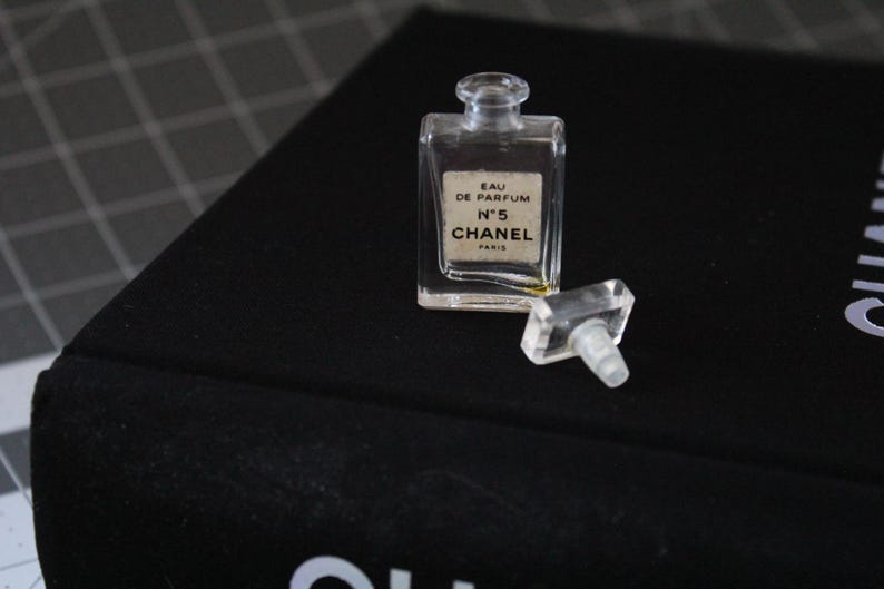 May include: A small, clear glass perfume bottle with a rectangular shape and a matching clear stopper. The label on the front reads EAU DE PARFUM N&deg;5 CHANEL PARIS in black text. The bottle is empty and sits upright with the stopper placed beside it.