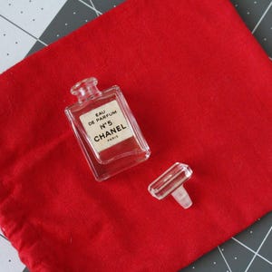 May include: A miniature clear glass perfume bottle with a rectangular shape and a detached clear stopper. The label reads EAU DE PARFUM N&deg;5 CHANEL PARIS. The bottle is placed on a bright red fabric drawstring pouch with a visible red cord.