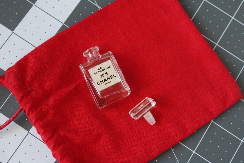 May include: A miniature clear glass perfume bottle with a rectangular shape and a detached clear stopper. The label reads EAU DE PARFUM N&deg;5 CHANEL PARIS. The bottle is placed on a bright red fabric drawstring pouch with a visible red cord.