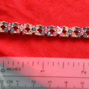 May include: A gold bracelet adorned with pink and purple gemstones. The bracelet is approximately 2.5 cm long. The gemstones are arranged in a repeating pattern. The bracelet is on a red surface.