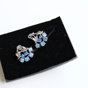 May include: A pair of silver earrings with blue and light blue rhinestones in a floral design.