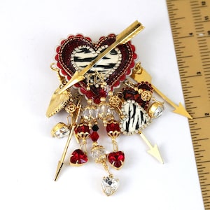 May include: A gold heart-shaped brooch with a zebra print design and red glitter. The brooch is adorned with multiple gold arrows, red and clear crystals, and a dangling heart charm.