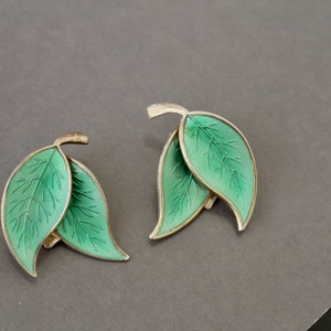Puede incluir: Un par de pendientes de plata con hojas de esmalte verde. Las hojas están detalladas con venas y tienen una forma ligeramente curvada.