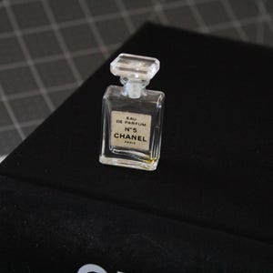May include: A miniature clear glass perfume bottle with a rectangular shape and a faceted clear stopper. The front label features the text EAU DE PARFUM N&deg;5 CHANEL PARIS in black lettering on a white background. The bottle contains a small amount of liquid.