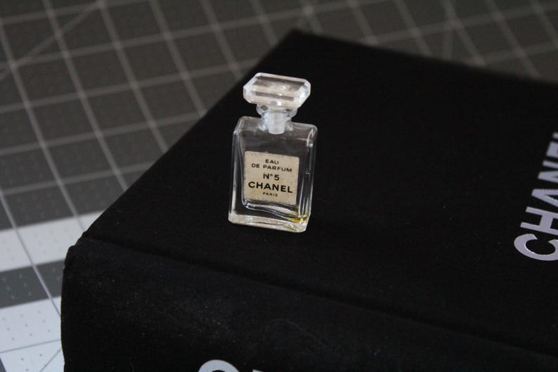 May include: A miniature clear glass perfume bottle with a rectangular shape and a faceted clear stopper. The front label features the text EAU DE PARFUM N&deg;5 CHANEL PARIS in black lettering on a white background. The bottle contains a small amount of liquid.