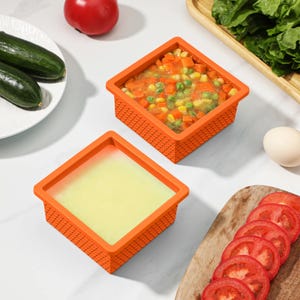 Op de afbeelding: Twee oranje vierkante voedselcontainers, één gevuld met gemengde groenten en de andere met een lichtgele substantie. Gesneden tomaten, komkommers en een tomaat zijn ook zichtbaar, wat duidt op voedselbereiding.