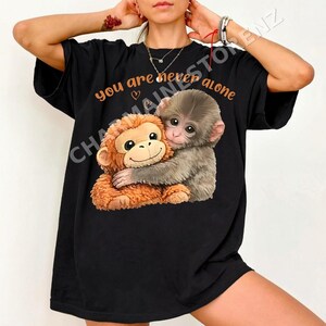 Puede incluir: Camiseta negra con un gráfico de dos monos abrazándose. El texto naranja "you are never alone" está impreso encima. La camiseta es de corte holgado.