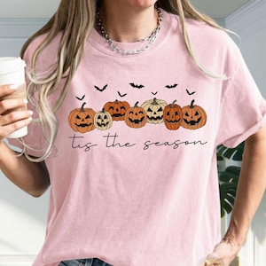 Puede incluir: Una camiseta de color rosa claro con un gr&aacute;fico de seis calabazas naranjas con caras talladas y varios murci&eacute;lagos negros peque&ntilde;os. Debajo de las calabazas, el texto 'tis the season' est&aacute; impreso en una fuente cursiva de color negro.