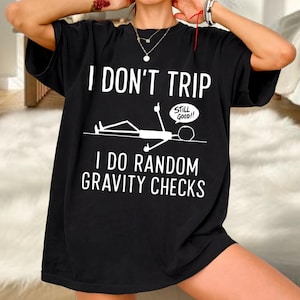 Può includere: Una t-shirt nera a maniche corte con testo bianco che recita I DON'T TRIP I DO RANDOM GRAVITY CHECKS. La grafica include un omino stilizzato bianco sdraiato a terra con un fumetto che riporta la scritta STILL GOOD!! in bianco.