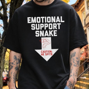 Op de afbeelding: Een zwart T-shirt met korte mouwen en de witte tekst "EMOTIONAL SUPPORT SNAKE" boven een witte pijl die naar beneden wijst. In de pijl staat een lijntekening van een slang met rode accenten en de rode tekst "CAUTION: HE SPITS".