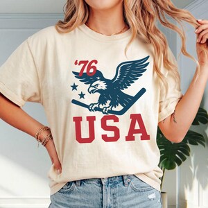 May include: A cream-colored short-sleeve t-shirt featuring a graphic print of a dark blue eagle holding a hockey stick. Above the eagle is the text '76' with two stars, and below the eagle is the red text 'USA' in a bold, block-style font.