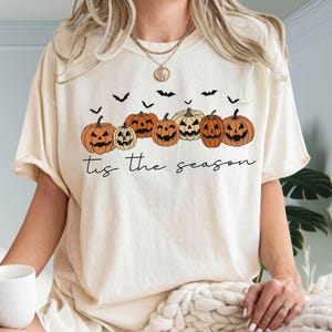 Puede incluir: Una camiseta de color crema con un gr&aacute;fico de seis calabazas de Halloween naranjas de varios tama&ntilde;os y formas, intercaladas con peque&ntilde;os murci&eacute;lagos negros. Debajo de las calabazas, el texto 'tis the season' est&aacute; impreso en cursiva negra.
