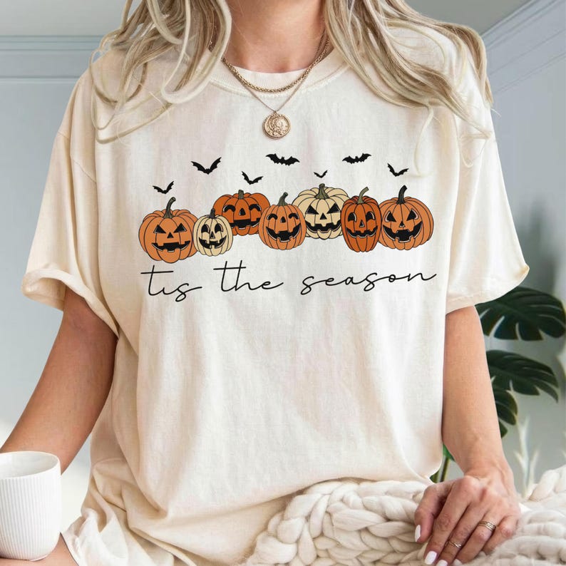 Puede incluir: Una camiseta de color crema con un gr&aacute;fico de seis calabazas de Halloween naranjas de varios tama&ntilde;os y formas, intercaladas con peque&ntilde;os murci&eacute;lagos negros. Debajo de las calabazas, el texto 'tis the season' est&aacute; impreso en cursiva negra.