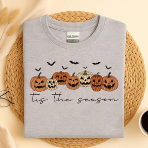 Puede incluir: Una camiseta de manga corta gris claro con una etiqueta Gildan Heavy Cotton. El gr&aacute;fico presenta una fila de siete calabazas de Halloween naranjas y crema con siluetas de murci&eacute;lagos negros y el texto 'tis the season' en letra negra.