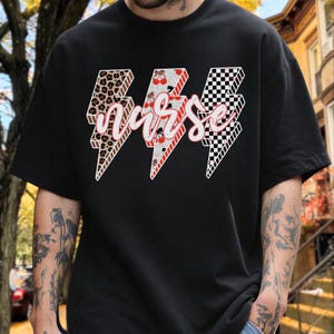 Peut inclure: Un t-shirt noir à manches courtes avec trois motifs d'éclairs sur la poitrine. Les éclairs présentent des imprimés léopard, des motifs de cerises et un damier noir et blanc. Le mot nurse est écrit en lettres cursives blanches par-dessus.