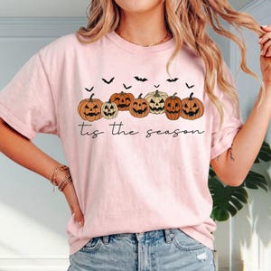 Puede incluir: Una camiseta de color rosa claro con un gr&aacute;fico de varias calabazas naranjas talladas como linternas y murci&eacute;lagos negros. Debajo de las calabazas, el texto 'tis the season' est&aacute; impreso en una tipograf&iacute;a cursiva de color negro.