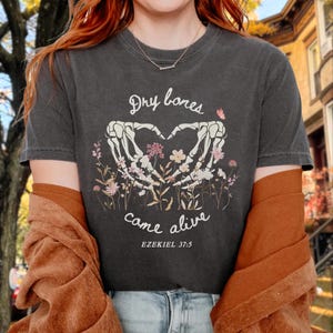 Könnte beinhalten: Ein anthrazitfarbenes T-Shirt mit einer Grafik aus zwei skelettierten Händen, die ein Herz über einem Blumenstrauß bilden. Der Text lautet Dry bones came alive und EZEKIEL 37:5 in weißer Schrift, ergänzt durch einen kleinen rosa Schmetterling.