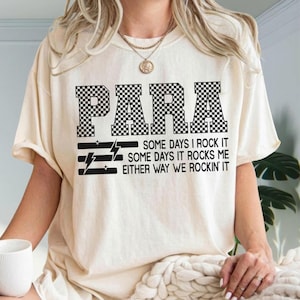 May include: A cream-colored t-shirt featuring the word PARA in a black and white checkered pattern. Below the text, the shirt displays the phrase SOME DAYS I ROCK IT SOME DAYS IT ROCKS ME EITHER WAY WE ROCKIN' IT with black lightning bolt icons.
