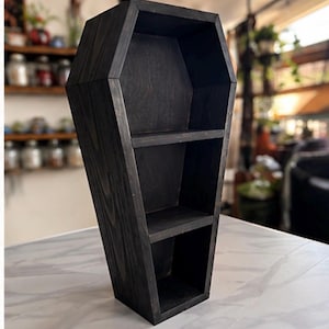 May include: A black wooden shelf shaped like a coffin with a hexagonal outline. The structure features two horizontal shelves creating three distinct storage compartments. The wood grain is visible through the dark stain on the exterior and interior.