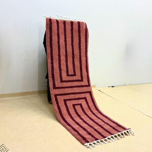 Puede incluir: Una alfombra rectangular con un diseño geométrico. La alfombra presenta un fondo rojo-naranja claro con patrones rectangulares marrón-rojizo oscuro. La alfombra tiene flecos en un extremo. La alfombra está hecha de un material suave y afelpado.