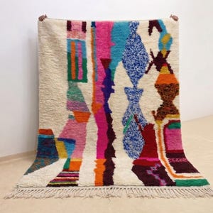 Puede incluir: Una alfombra rectangular con base de color crema que presenta un patrón abstracto vibrante y multicolor. El diseño incluye rayas verticales y formas geométricas en tonos rosa intenso, naranja, azul, verde y morado oscuro, con flecos.