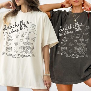 K&ouml;nnte beinhalten: Zwei &uuml;bergro&szlig;e T-Shirts, eines in Elfenbein und eines in Pfeffer, mit einer schwarz-wei&szlig;en Grafik. Die Grafik enth&auml;lt den Text "Seashells & Wedding Bells" und "Kathleen's Bachelorette, Cape Cod, MA" sowie Illustrationen von Muscheln, Sektgl&auml;sern und Glocken.