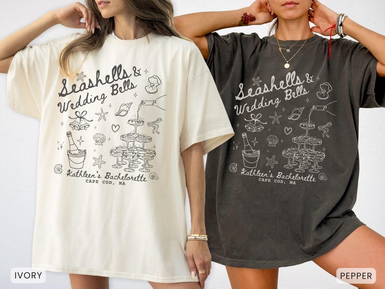 K&ouml;nnte beinhalten: Zwei &uuml;bergro&szlig;e T-Shirts, eines in Elfenbein und eines in Pfeffer, mit einer schwarz-wei&szlig;en Grafik. Die Grafik enth&auml;lt den Text "Seashells & Wedding Bells" und "Kathleen's Bachelorette, Cape Cod, MA" sowie Illustrationen von Muscheln, Sektgl&auml;sern und Glocken.