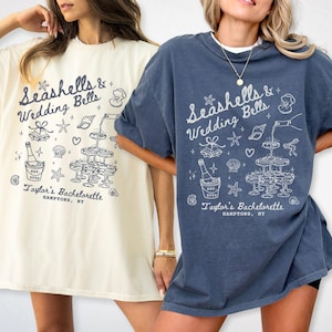 May include: Two t-shirts, one ivory and one denim, featuring a white graphic. The design includes the text "Seashells & Wedding Bells" and "Taylor's Bachelorette, Hamptons, NY" with illustrations of champagne and seashells.