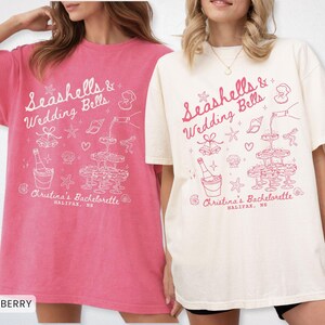 K&ouml;nnte beinhalten: Zwei &uuml;bergro&szlig;e T-Shirts, eines in Crunchberry und das andere in Elfenbein, zeigen ein wei&szlig;es Grafikdesign. Das Design enth&auml;lt den Text "Seashells & Wedding Bells" und Illustrationen von Champagner, Muscheln und einem Champagnerturm. Der Text "Christina's Bachelorette, Halifax, NS" ist ebenfalls enthalten.