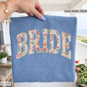 May include: Blue jean sweatshirt with the word "BRIDE" in a floral design. The letters are filled with pink, orange, and green flowers on a striped background. The text "FAUX PEARL DESIGN" is above the sweatshirt, and "BLUE JEAN" is below.