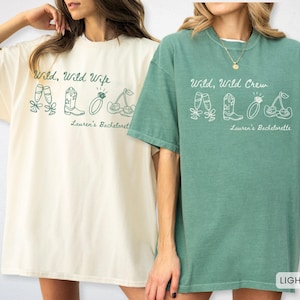 Puede incluir: Dos camisetas de gran tama&ntilde;o, una marfil y otra verde claro, con texto gr&aacute;fico blanco e ilustraciones. La camiseta marfil dice "Wild, Wild Wife" y la camiseta verde dice "Wild, Wild Crew". Ambas camisetas tienen "Lauren's Bachelorette" impreso debajo.